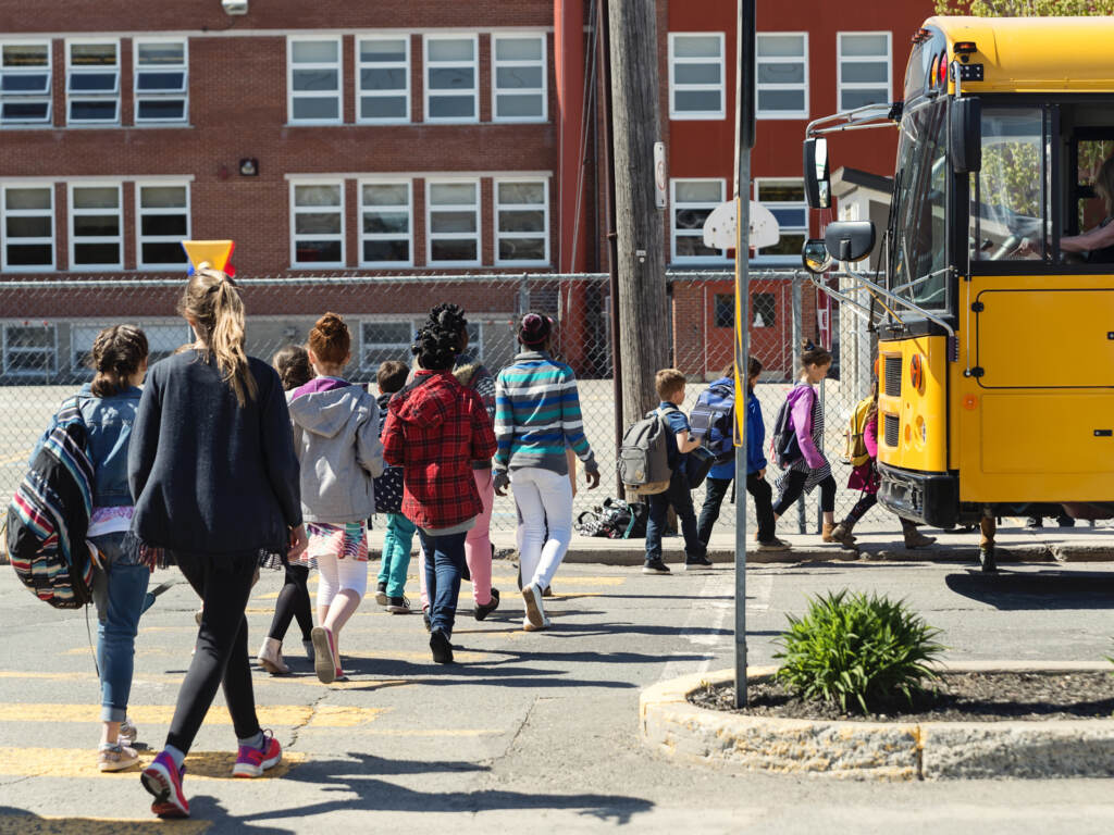 As kids return to school this fall, educators are prepared to deal with the continued mental health fallout of the disruptions of the pandemic. Students walk by a yellow school bus towards a red-brick school building.