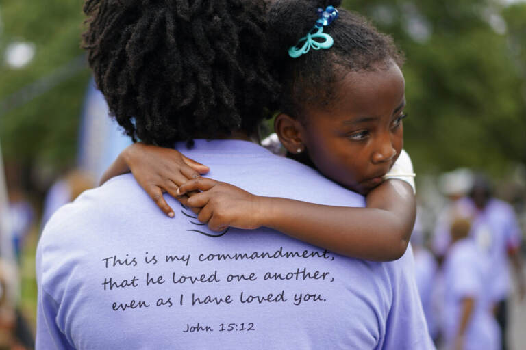 A little girl looks over the shoulder of an adult wearing a shirt which has a Bible verse written on the back.