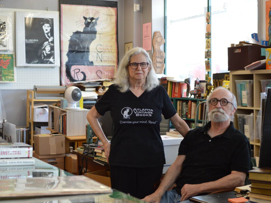 independent-bookstores-banned-books Two people, one sitting, one standing, look towards the camera, with bookshelves full of books visible behind them.