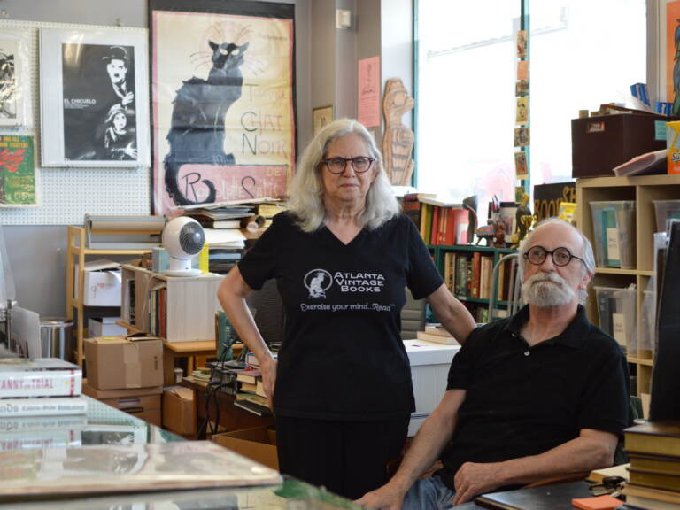 Two people, one sitting, one standing, look towards the camera, with bookshelves full of books visible behind them.
