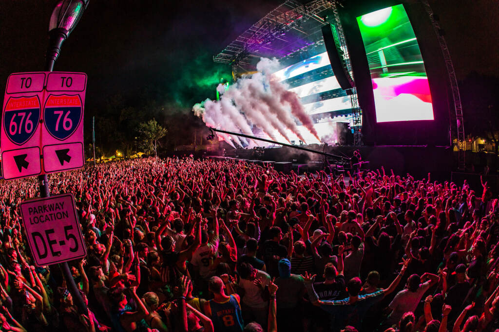 A crowd of people in front of a stage is seen from above, lit up by a pink light.