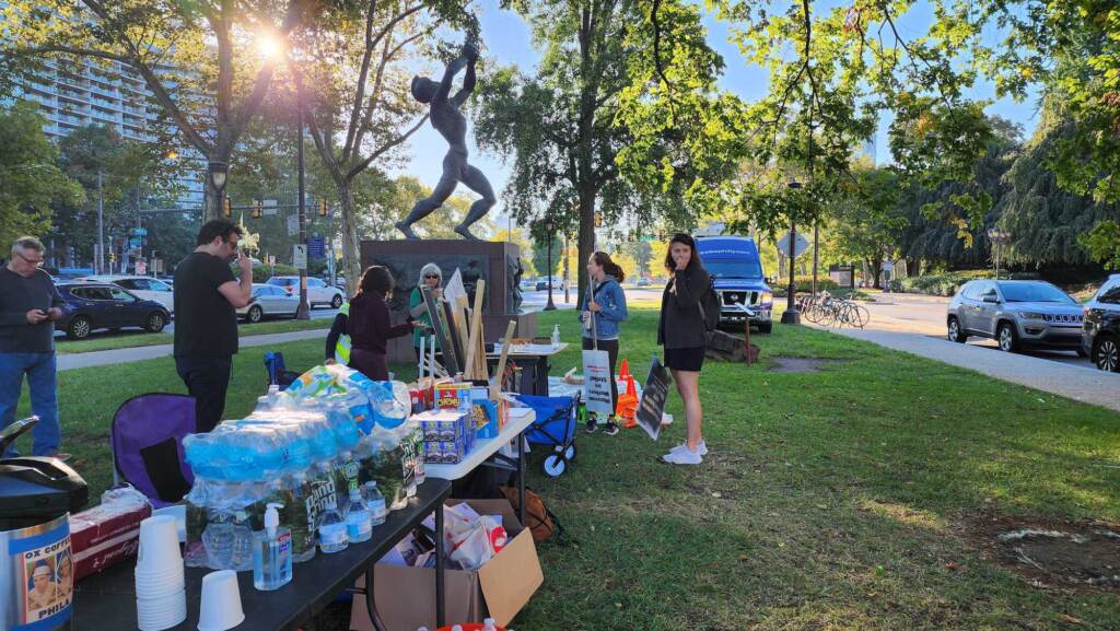philadelphia-art-museum-strike-1 People gather around a table with water bottles and snacks. A sculpture, trees, and street are visible in the background.