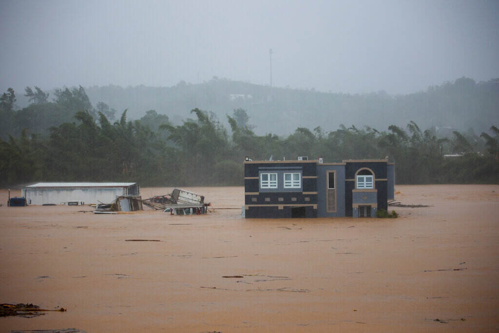 A home is surrounded by brown floodwaters as trees bend in the wind in the background.