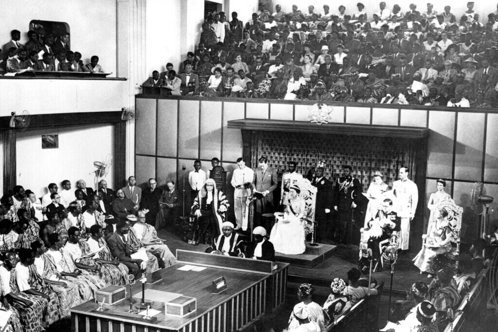 Britain Queen Colonialism’s Shadow A black and white photo shows the Queen in a large room filled with people.