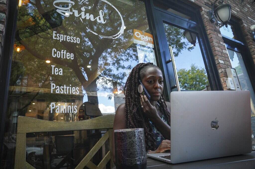 Kymme Williams-Davis A woman holds a phone up to her ear while looking at a laptop screen outside of a storefront.
