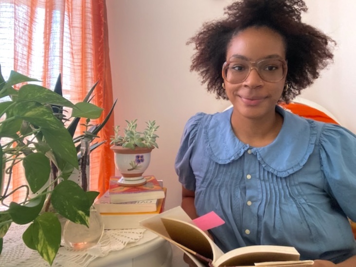 A woman smiles next to a desk, as she holds an open book.