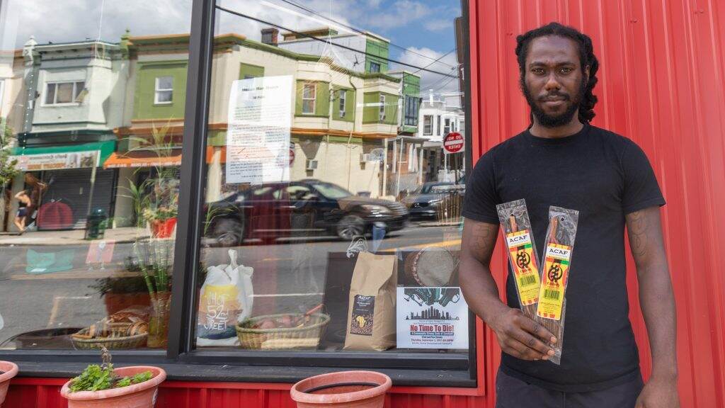 westphilly-52ndstreet-businessowners06-crop-1024×576 A man stands outside of a store, with the reflection of other stores on the street visible in the storefront window.
