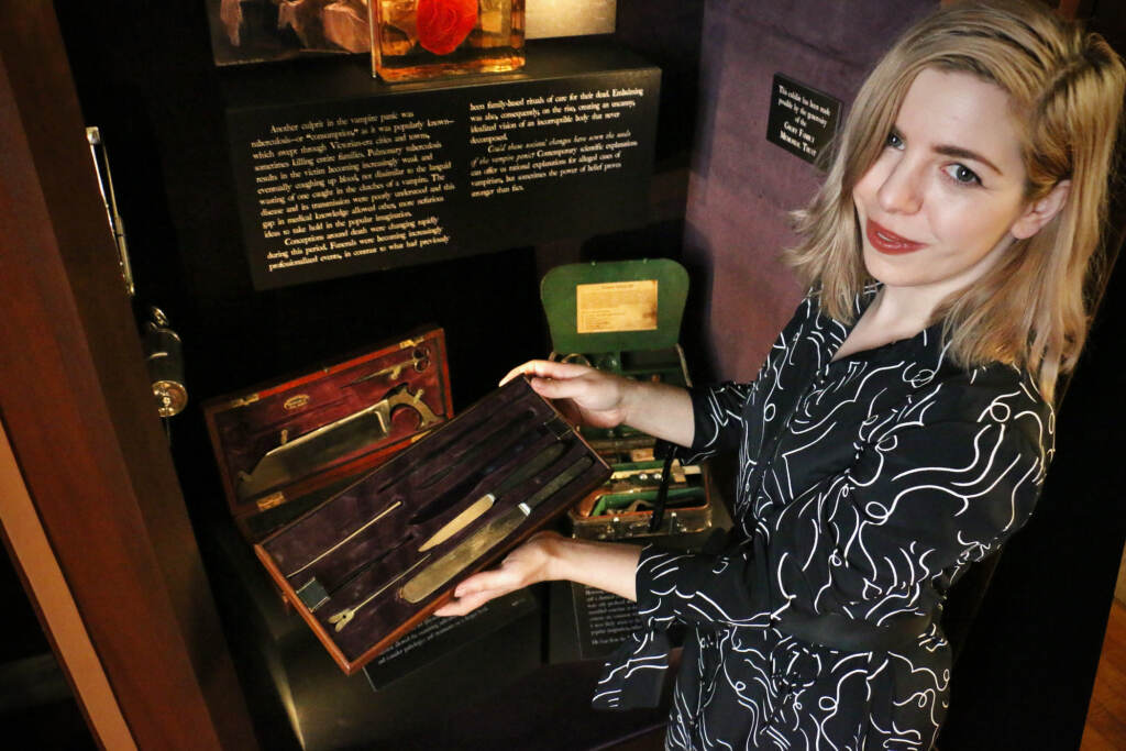 A woman displays a case of surgical instruments in front of an exhibit.