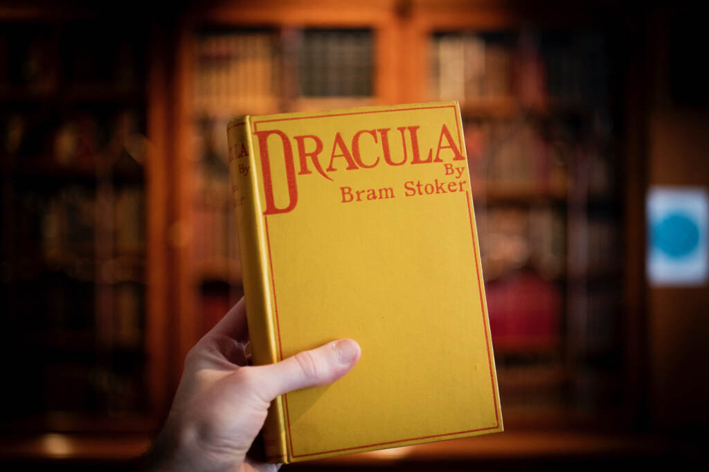 A hand holds up a yellow book with the title displayed in red, "Dracula," and the words "by Bram Stoker." Bookcases are visible in the background.