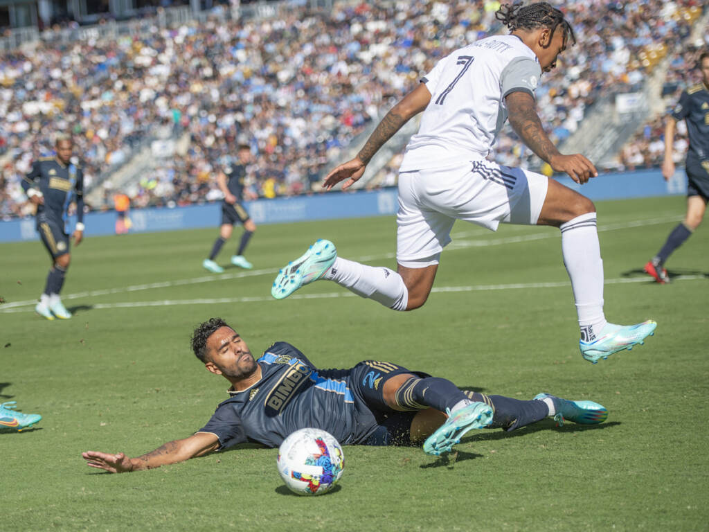 A soccer player is on the ground near the ball as a player on the opposing team jumps over him.