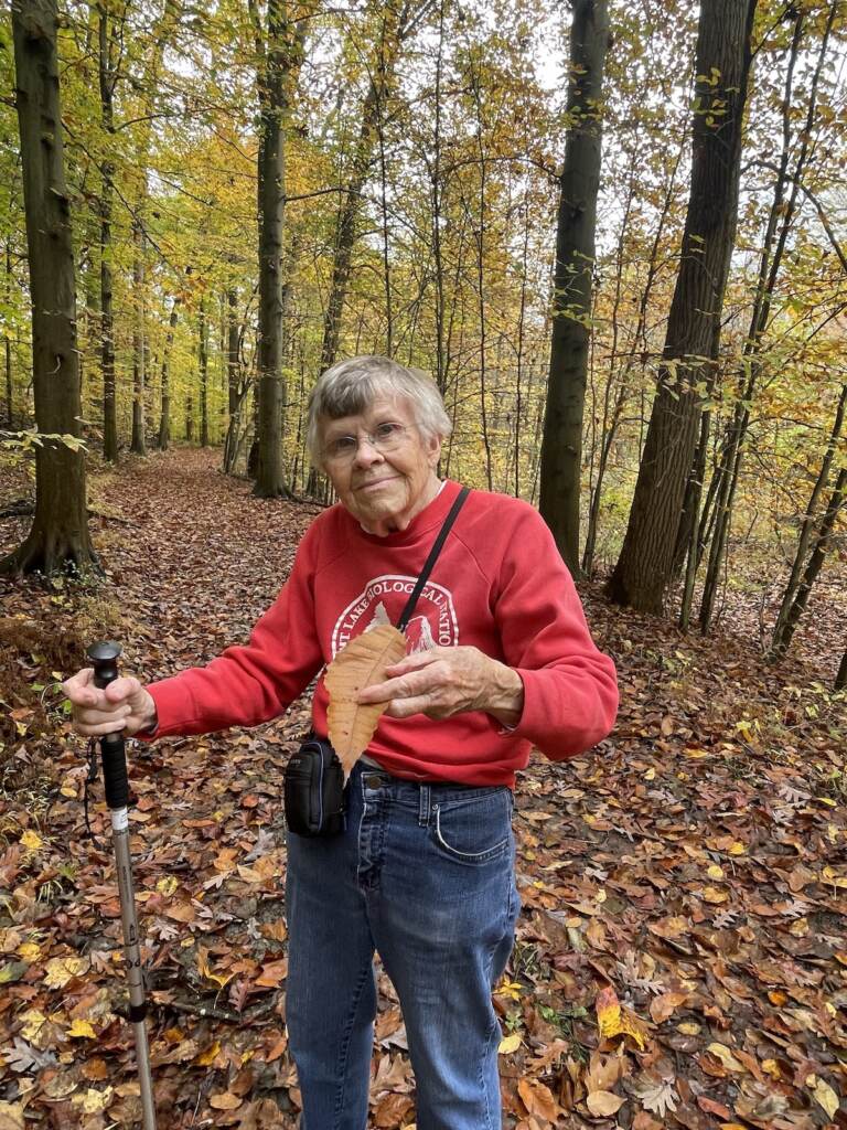 A woman holding a walking stick holds up a leaf with her other hand, as trees are visible behind her.