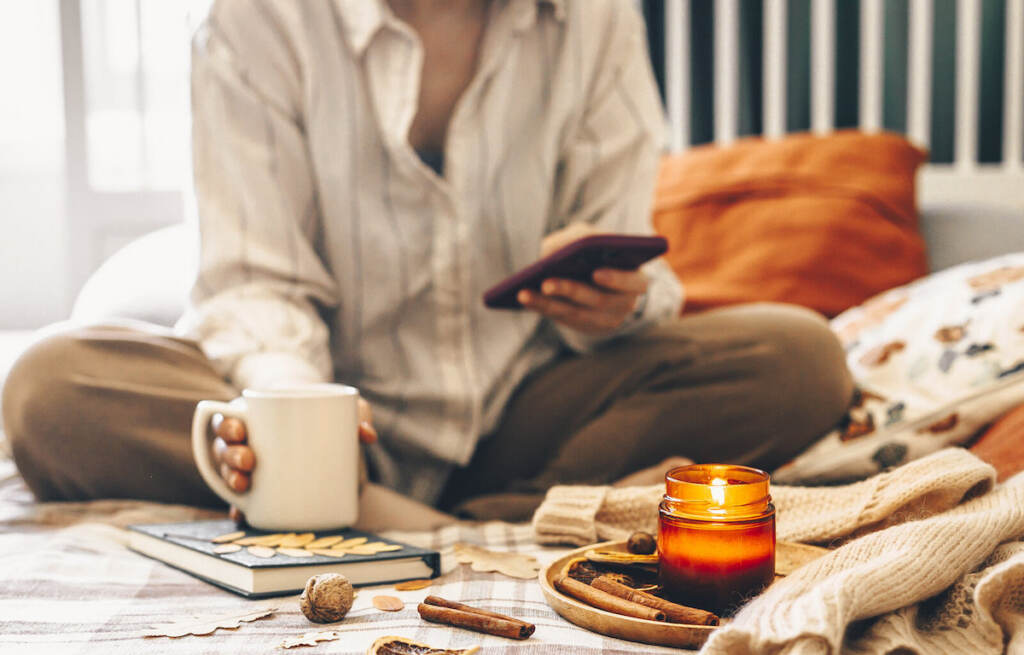 Woman sitting on bed in lotus pose using phone and drinking morrning beverage with burning candle An up-close view of a person sitting in bed, holding a coffee mug, with a candle lit in front of them.