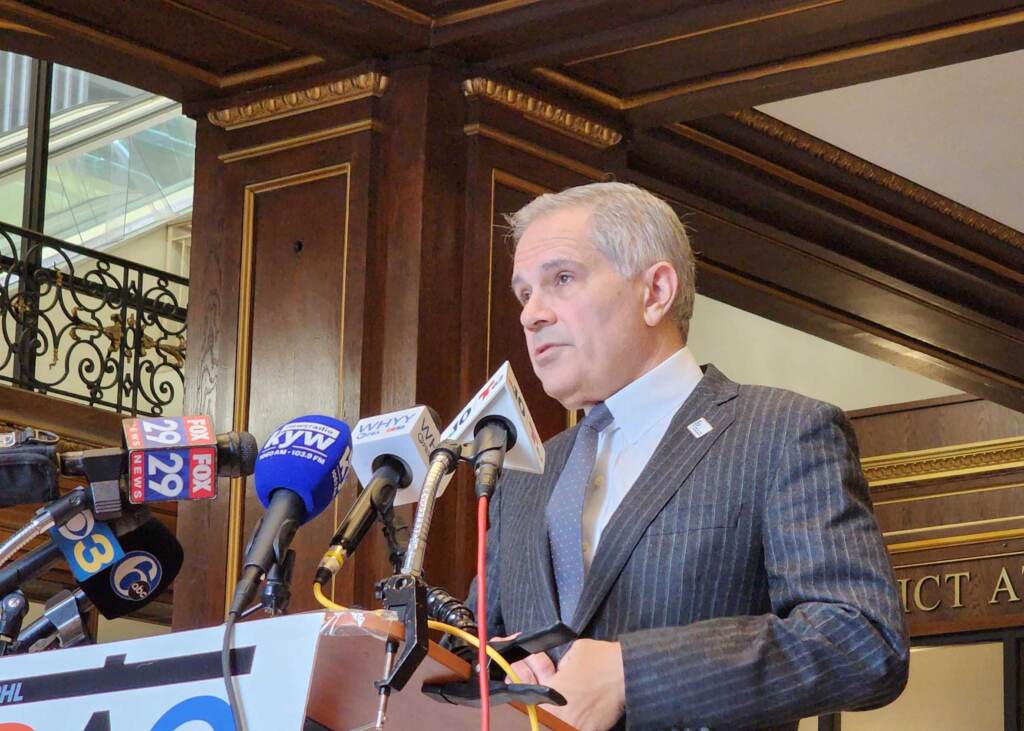 Krasner stands at a podium and speaks into microphones from various media outlets.