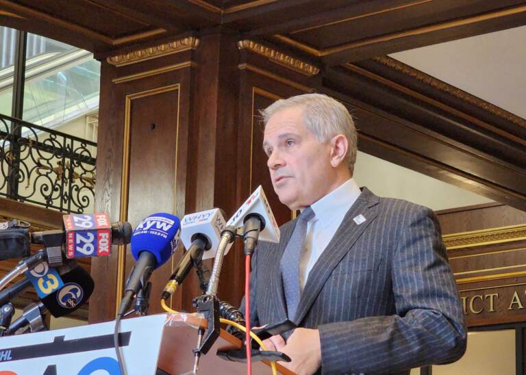Krasner stands at a podium and speaks into microphones from various media outlets.