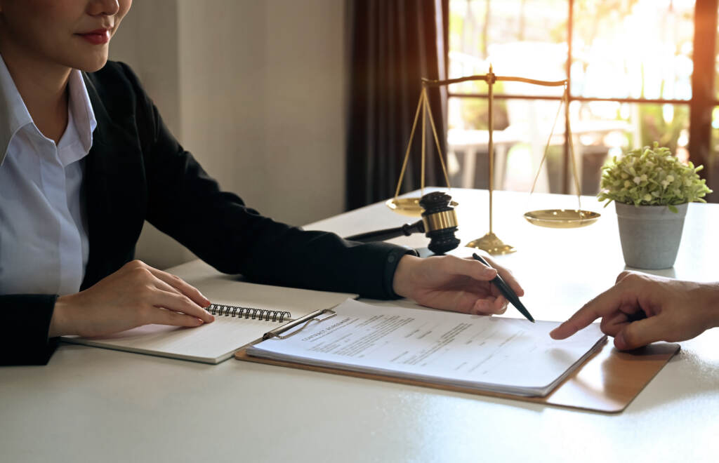 Female Lawyer Discussing Contract Document, Providing Law Consultation And Legal Advice To Her Clien A lawyer points to something on a document. The hand of the person seated across the table from her is visible.
