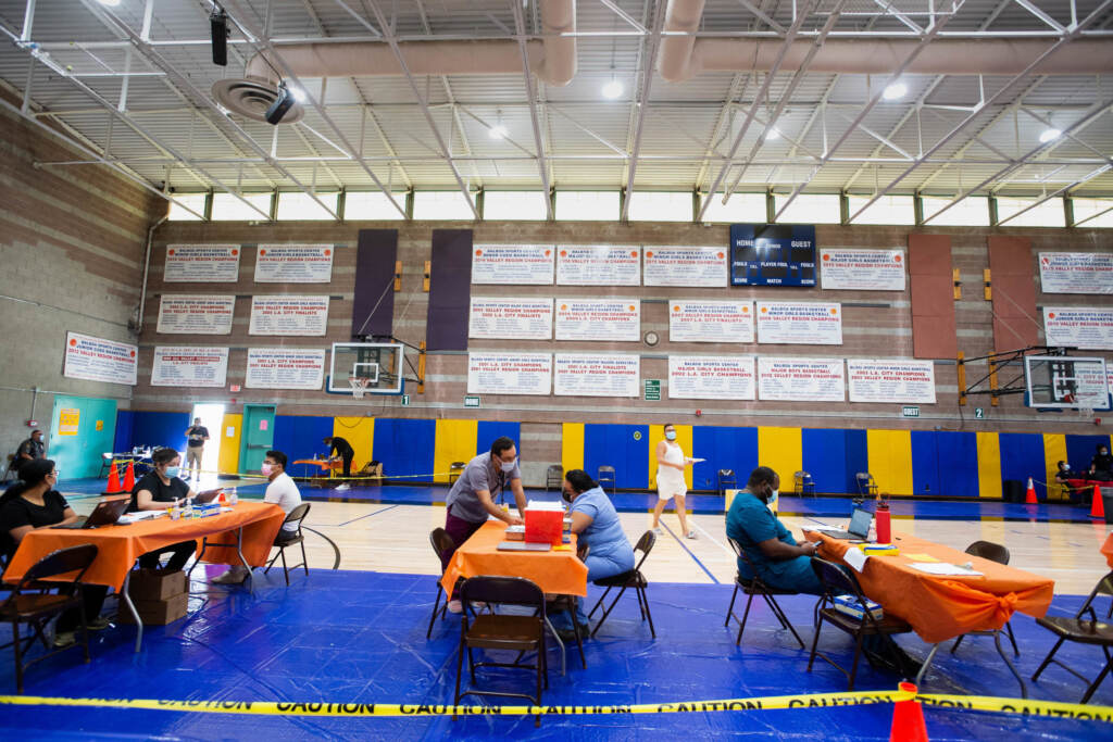 The new bivalent COVID-19 booster is offered by the Los Angeles County Department of Public Health. Health experts say getting more people boosted could help stave off a winter COVID surge. People are at tables in a vaccination clinic.