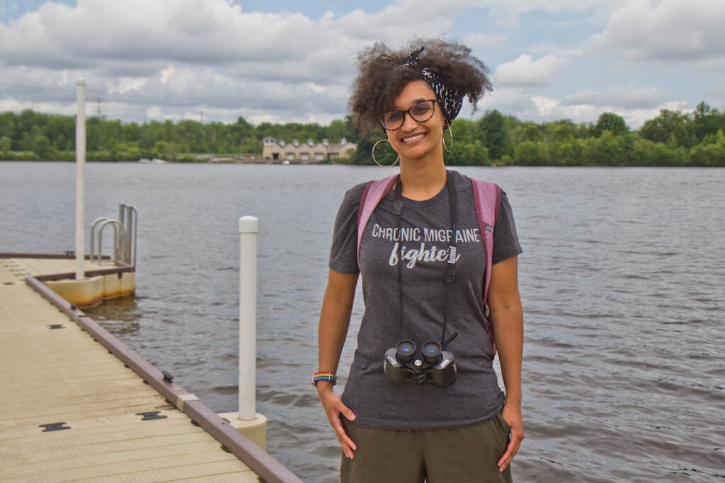 A woman stands on a bridge. Water and trees are visible behind her.