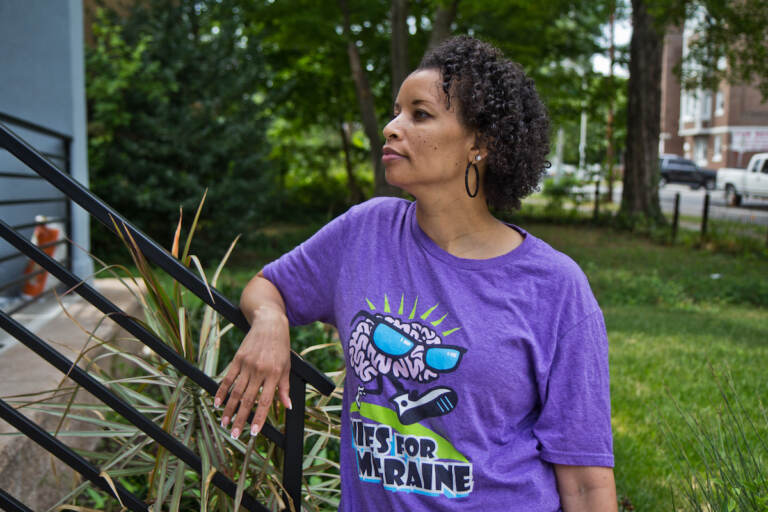 A woman gazes to the right. Trees and a street are visible behind her.