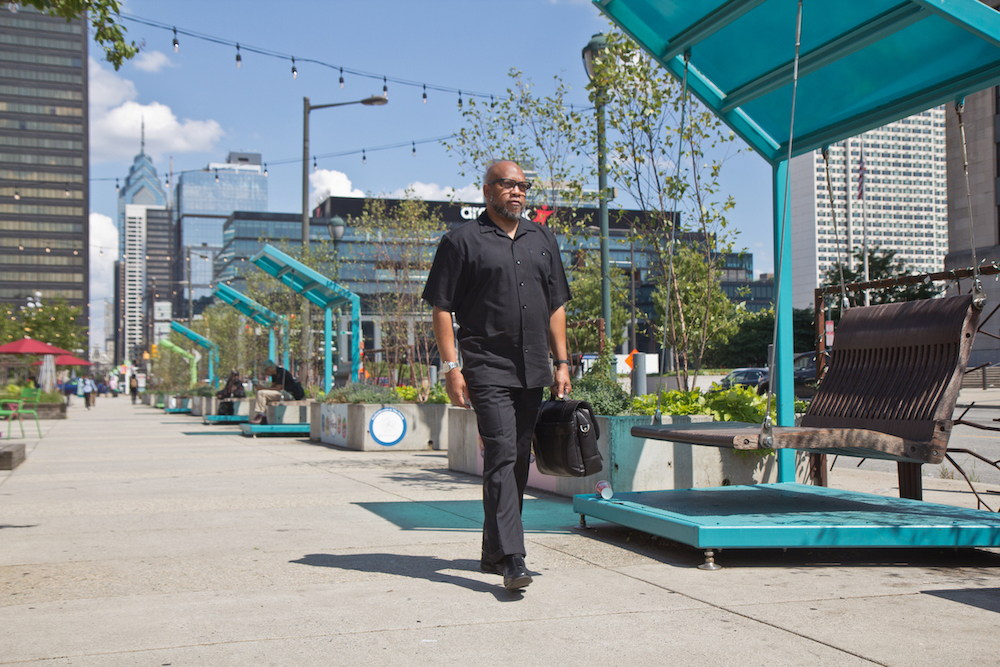 A man walks down the street with a briefcase. The Philadelphia skyline is visible behind him.