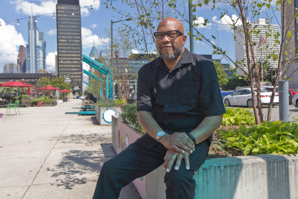 A man sits on a garden planter with the Philadelphia skyline visible behind him.
