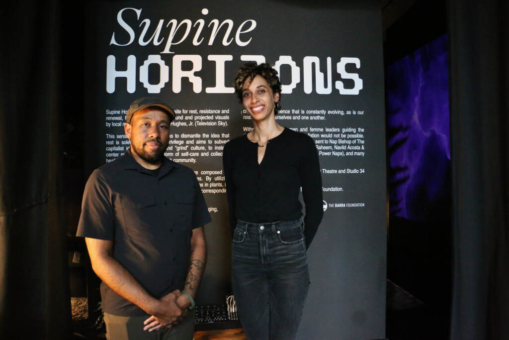 Two people stand in front of a sign that reads, "Supine Horizons."