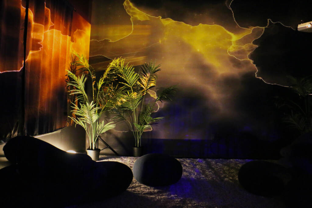 Orange and yellow lights flow over darkened walls. A few plants are lit up from a bright white light placed below them.