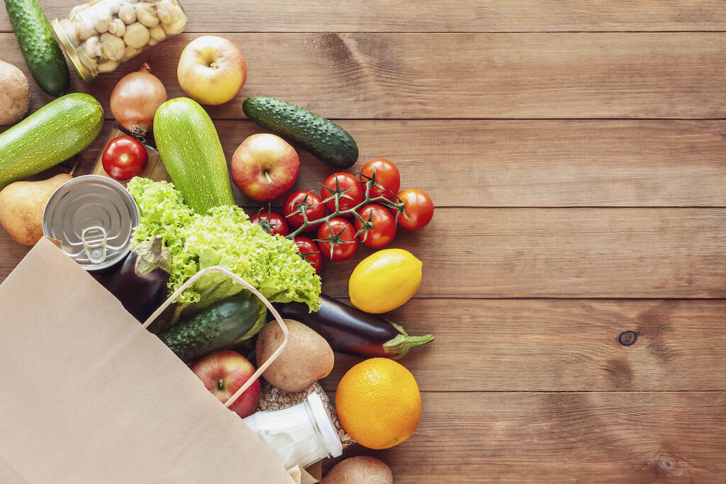 Paper Grocery Bag With Fresh Vegetables, Fruits, Milk And Canned Goods On Wooden Backdrop. Food Deli A bag of groceries is seen from above.
