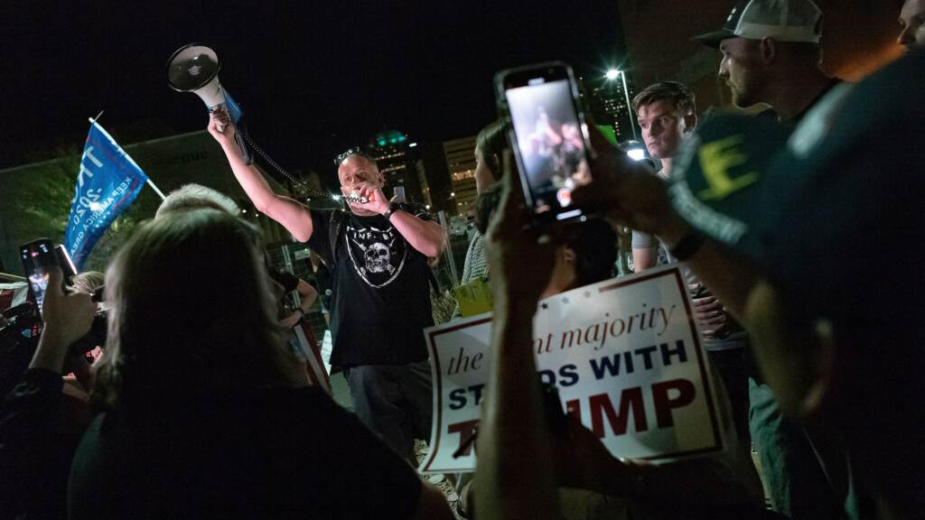 A man shouts into a bullhorn as Trump supporters gather in front of the Maricopa County Elections Department in Phoenix, where ballots were being counted following the 2020 presidential election. Federal officials have warned about violence erupting after this year’s election, with experts particularly concerned about the period after Nov. 8. A man yells into a bullhorn surrounded by other people.