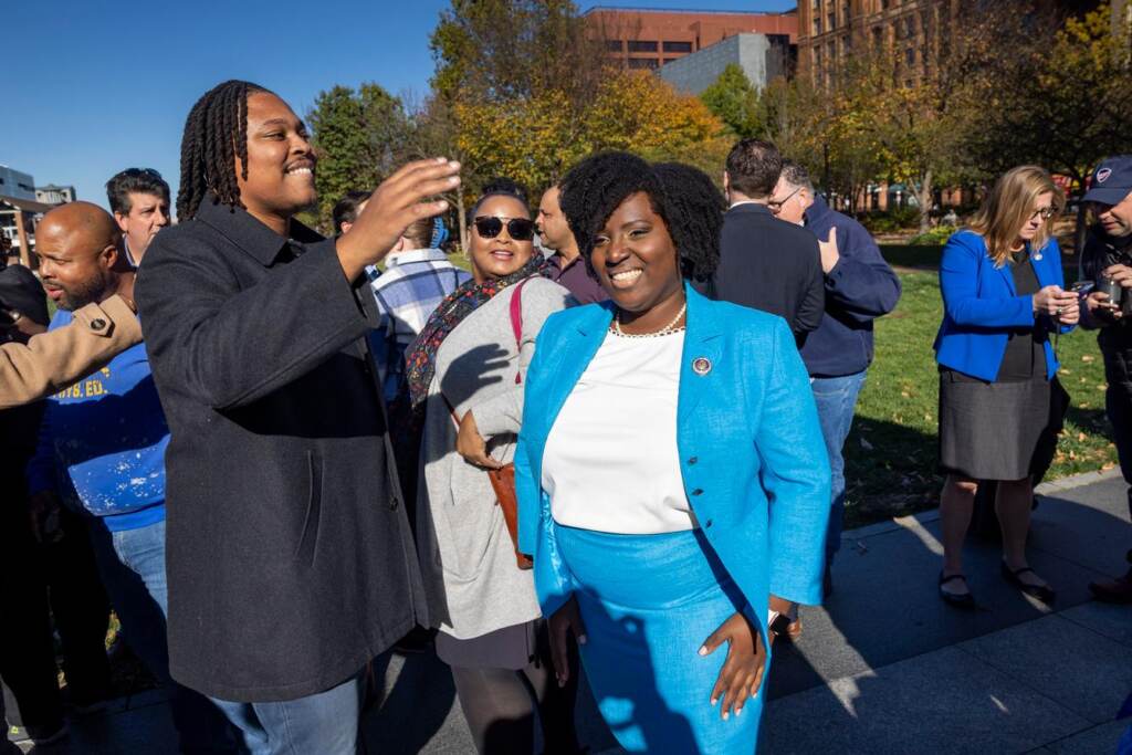 jt9d0ye6xqge0k55a40a7q0hm4 Rep. McClinton, wearing a bright blue suit, smiles in the midst of other legislators on a sunny day.