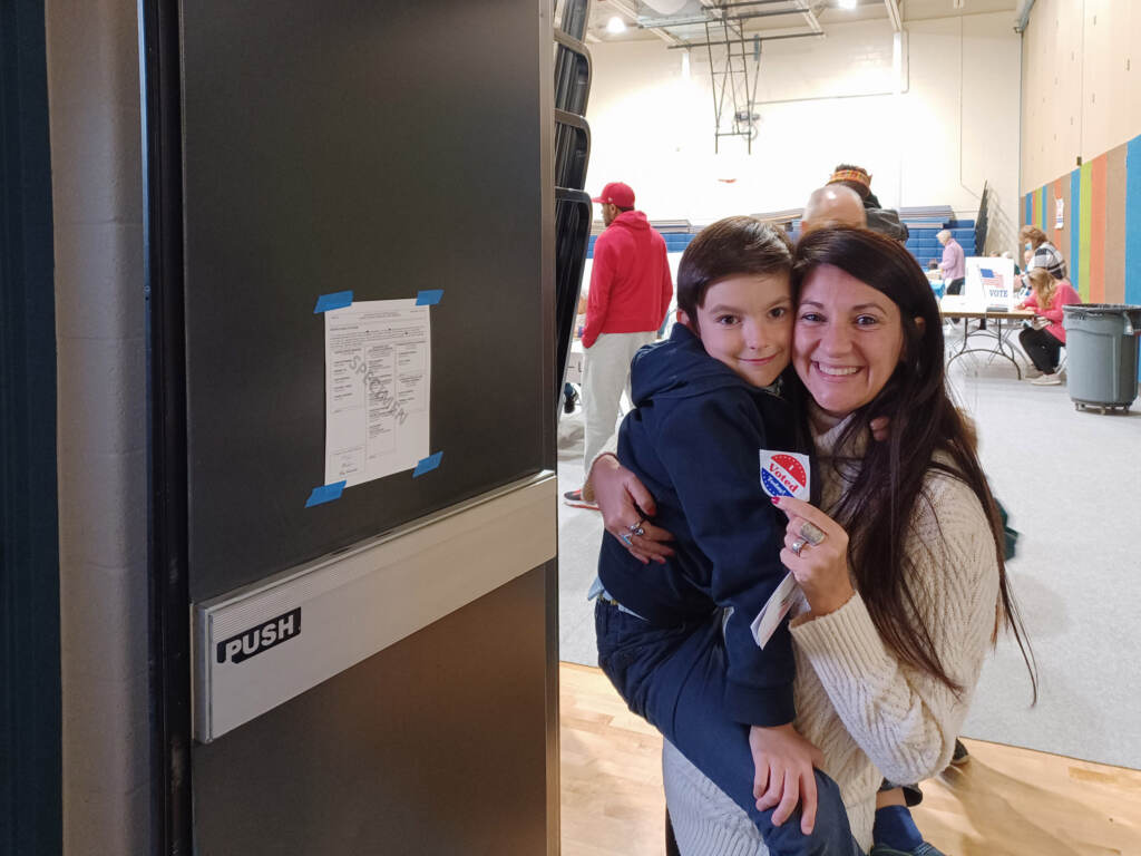A woman holds up an I Voted sticker while also holding her child.