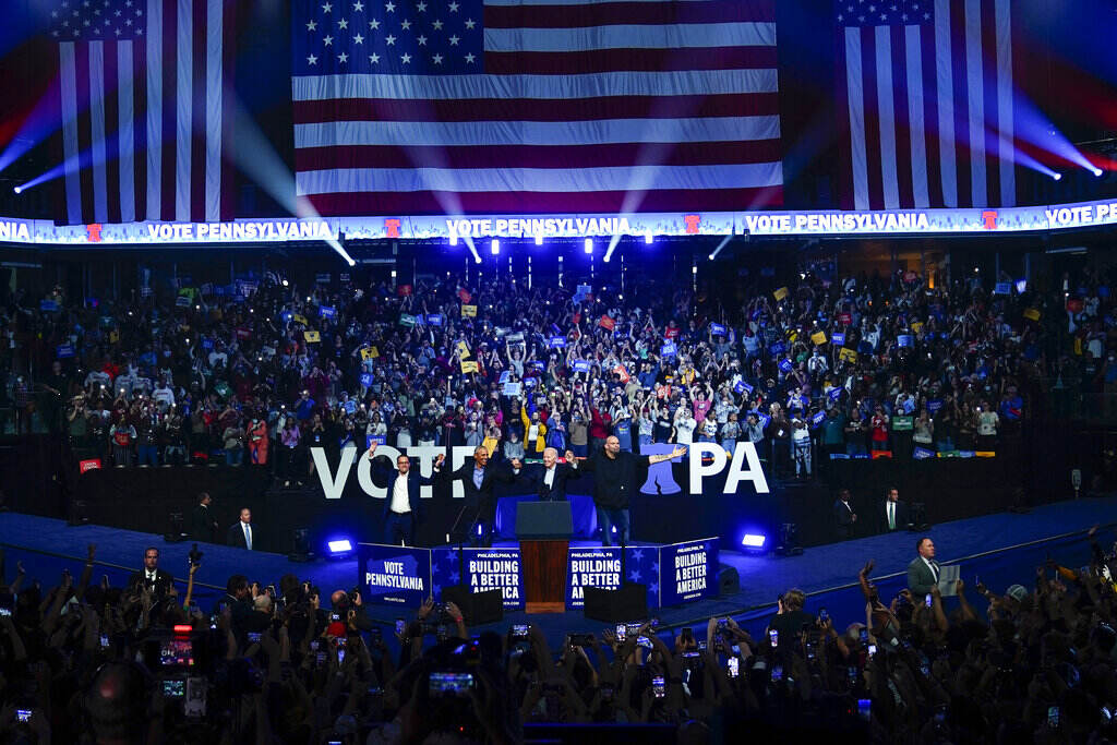 Shapiro, Obama, Biden, and Fetterman hold hands in front of a cheering crowd with an American flag displayed in the background.