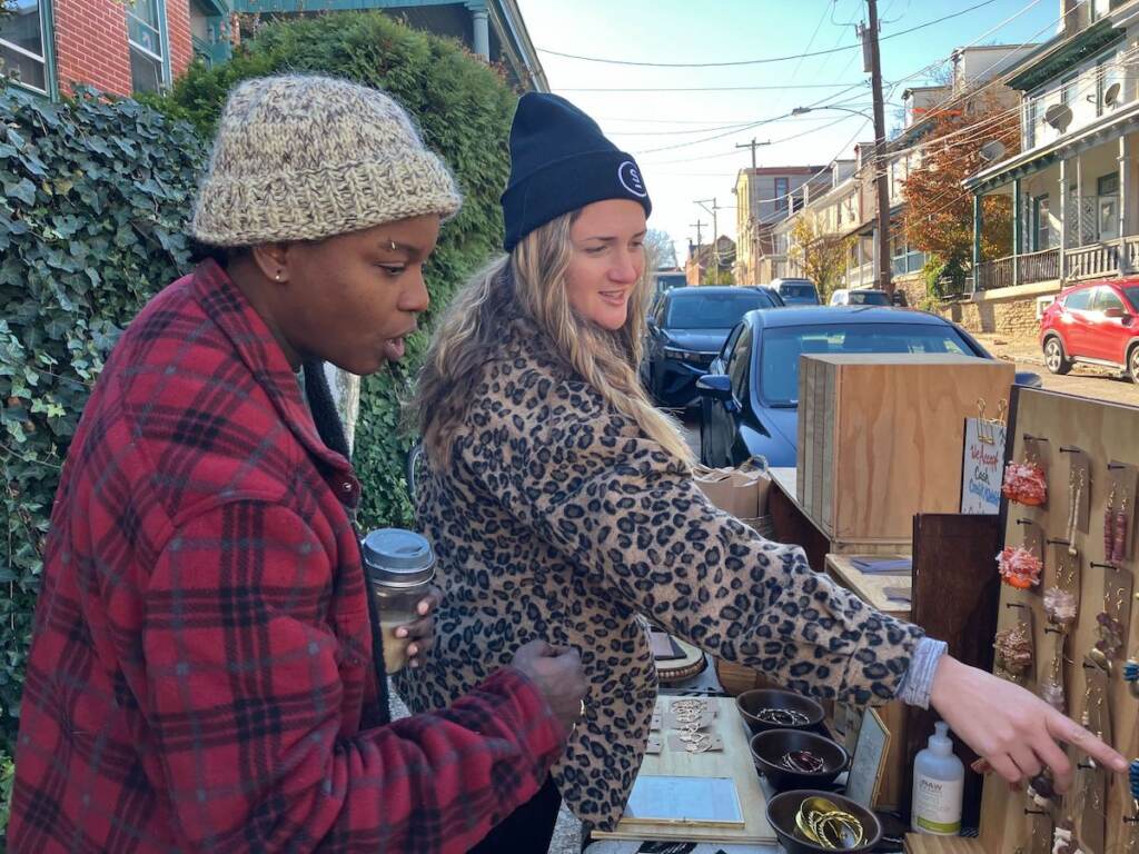Two people look at jewelry outdoors on a street.