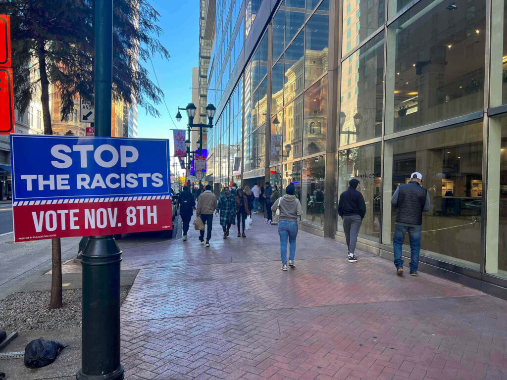 A sign reads Stop the racists as people walk on a sidewalk in the background.