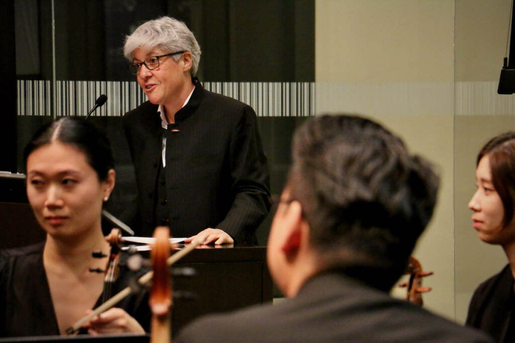 A woman speaks at a podium, with orchestra musicians visible in the foreground.