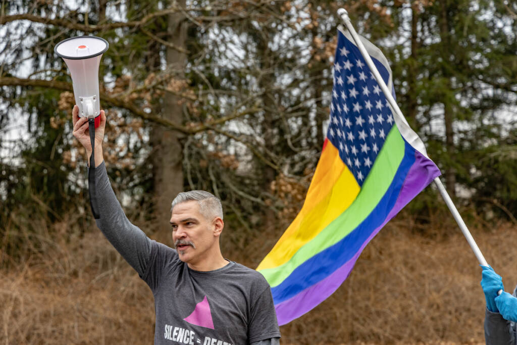 A person holds up a megaphone. A Pride version of the U.S. flag is visible in the background.