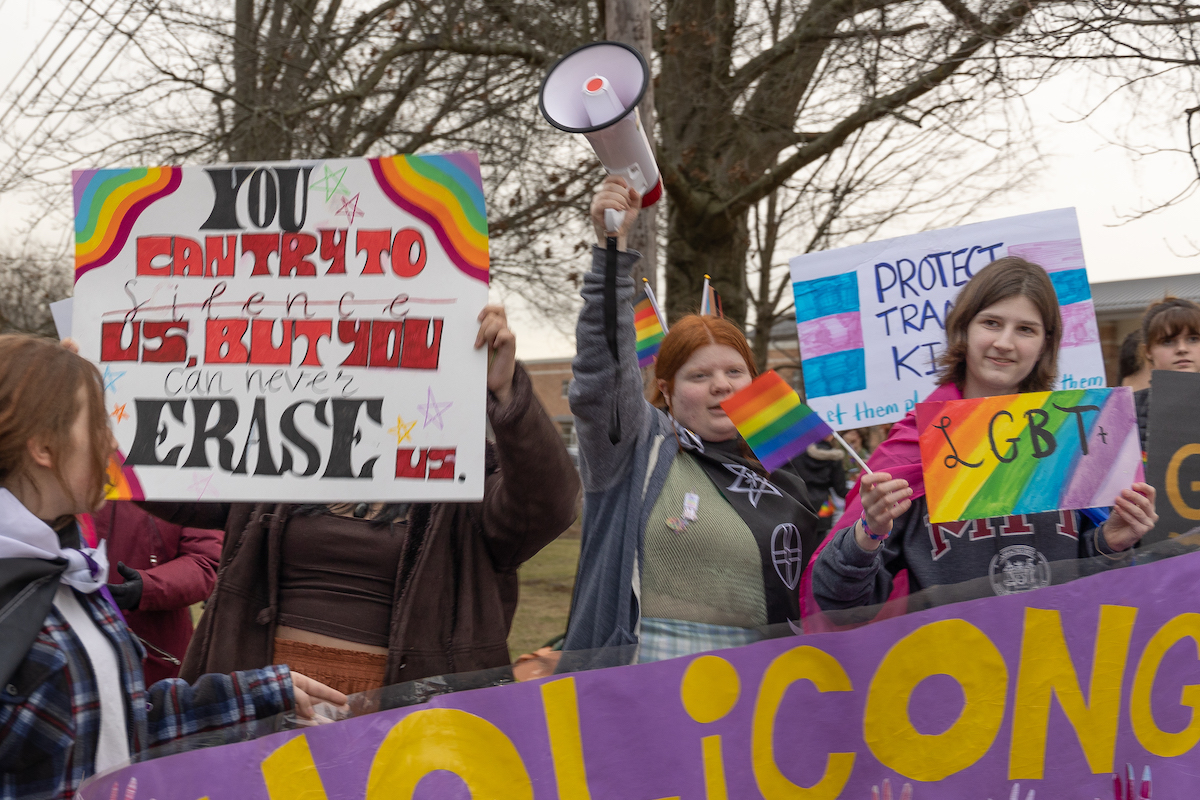 A group of students hold up Pride flags, a megaphone, and signs supporting LGBTQ students.