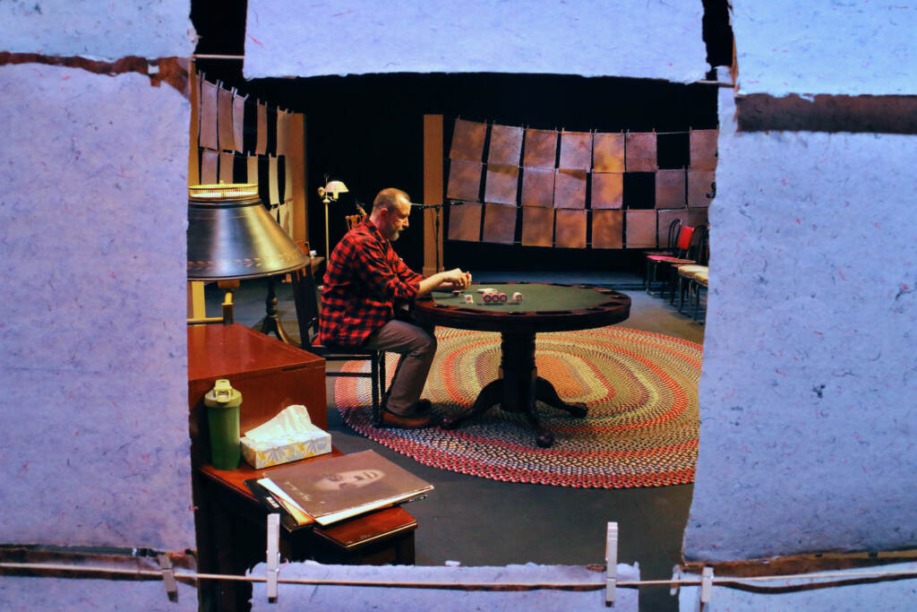2023 01 11-e lee-charlie delmarcelle-philadelphia louis bluver theatre at the drake-shadow that broke the light paper A person is seen through a gap in a row of papers strung on clotheslines. He's seated at a table, looking downwards.