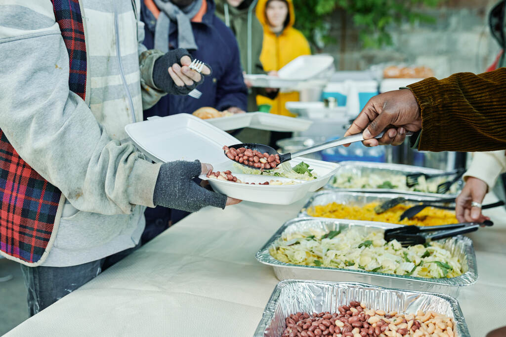 Close-up Of Volunteers Serving Homeless People And Putting Meal A person spoons food onto someone else's plate.