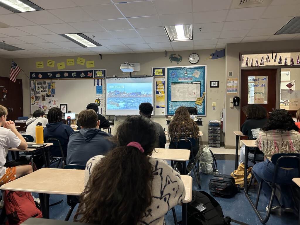 As seen from the back of the classroom, a group of students seated at desks look toward the front of the room where a screen is.