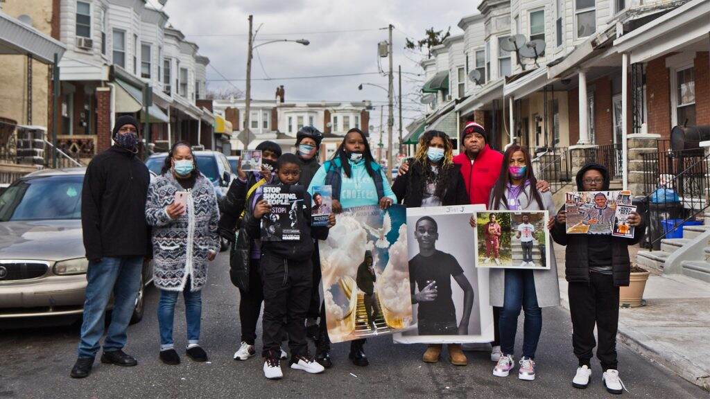 gunviolence-families-mlk-westphilly-crop-1024×576 A group of people stand together in the middle of a street holding photographs.