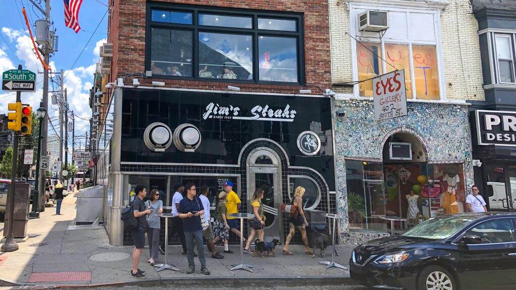 jimssteaks-eyesgallery-southstreet-crop-1024×576 People gather around Jim's Steaks storefront.