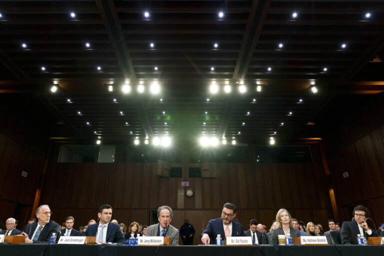 A row of people sit at microphones in the U.S. Senate chambers.