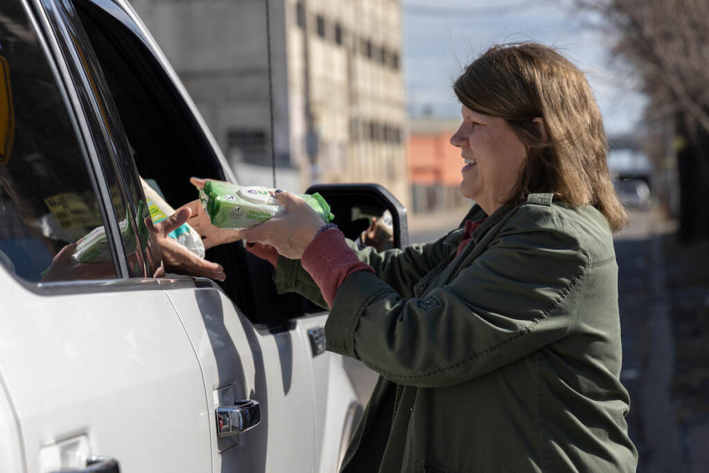 A woman hands diapers and other supplies to a person in the passenger side of a vehicle.