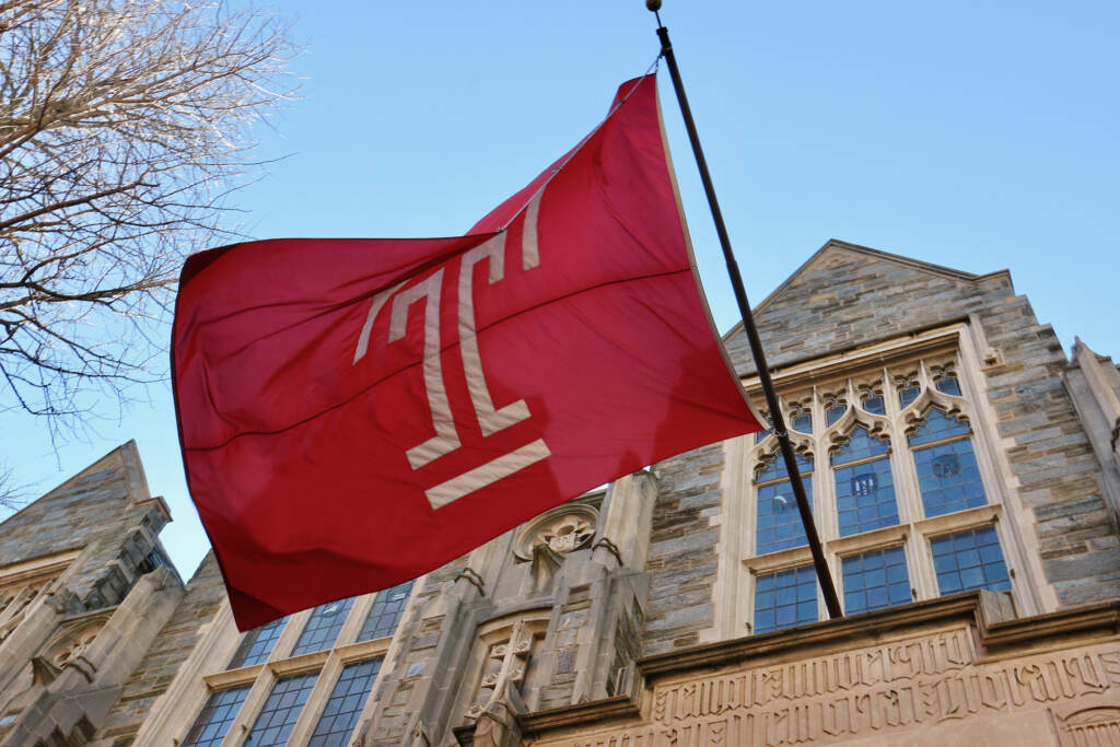 A flag flies on Temple University’s Sullivan Hall. (Emma Lee/WHYY) A view of a Temple flag from below, flying outside of a university building.