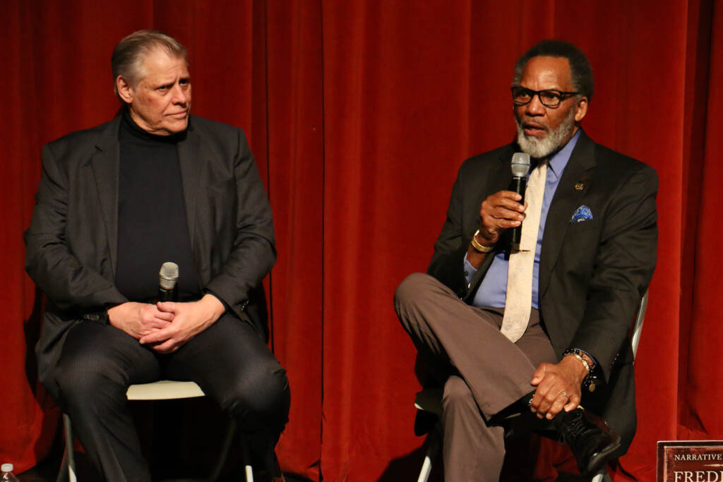 Two men speak onstage. Behind them is a bright red curtain.