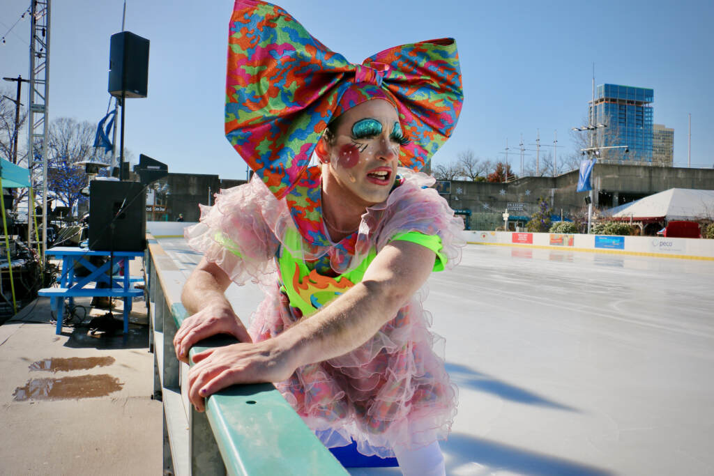 2023 02 18-e lee-john jarboe-philadelphia riverrink-beards on ice An ice skater and performer clutches the side of the ice skating rink on a sunny day.