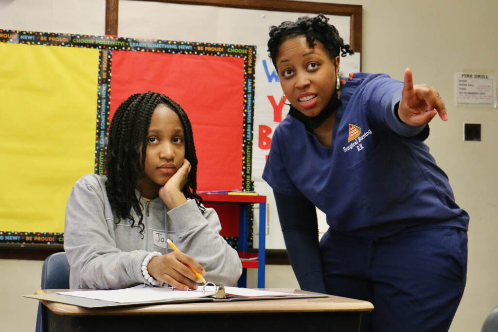 2023 02 21-e lee-jada whipple jamel mann-philadelphia george meade school-mini nurse academy A nurse stands next to a student seated at the desk.