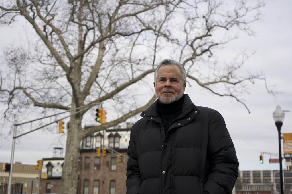 A man poses for the camera. A tree, buildings, and traffic lights are visible behind him.