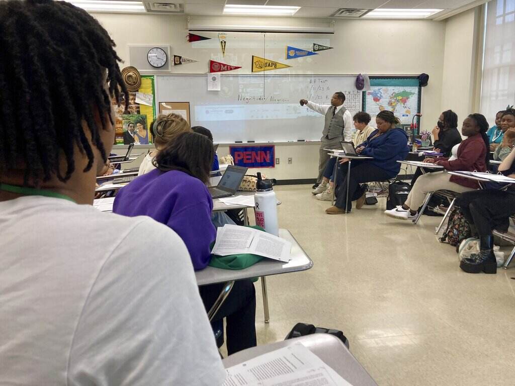 Students look towards the front of the room where their teacher is gesturing towards something written on the whiteboard.