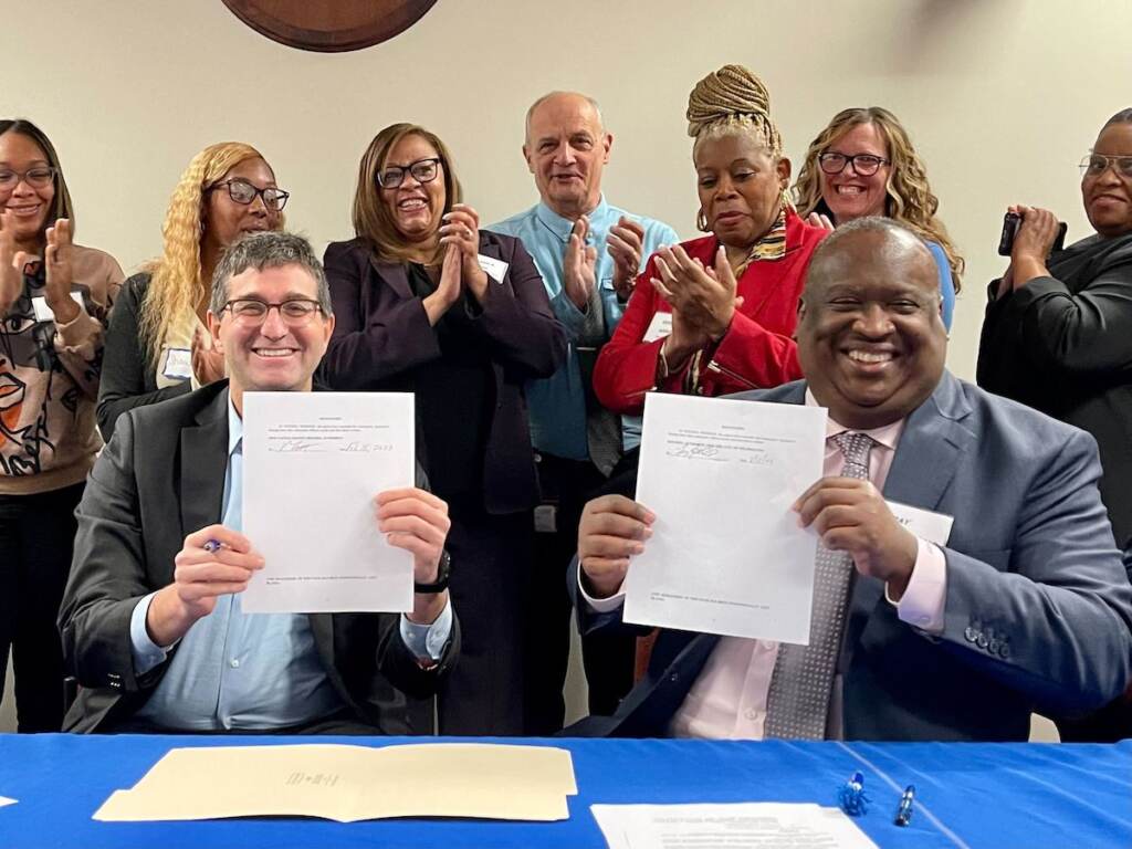 delaware-housing-vouchers-new-program Two people hold up signed documents while seated at a table. Other people pose for a photo in the background.